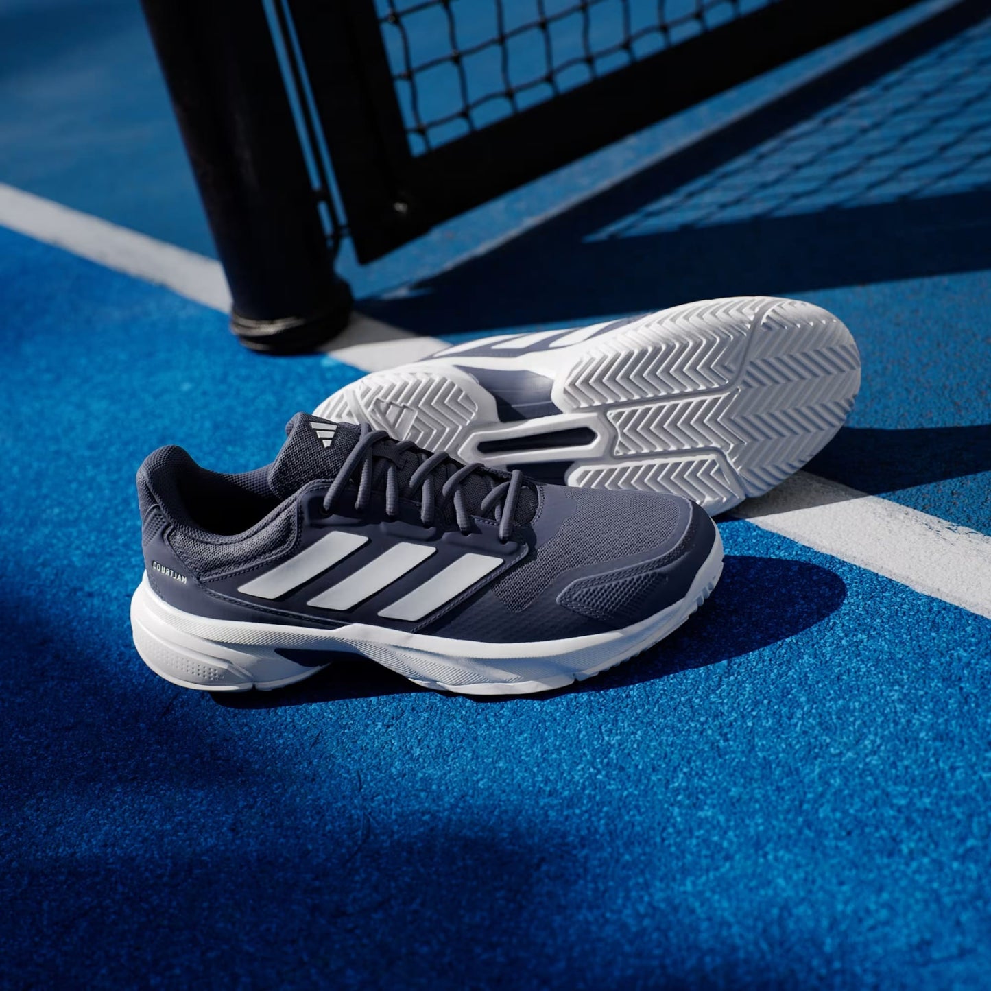 A pair of adidas CourtJam Control 3 Men’s Tennis Shoes in Dark Blue and Cloud White rests on a vibrant blue sports court near a black fence post and white boundary line, combining stylish design with reliable stability for every match.