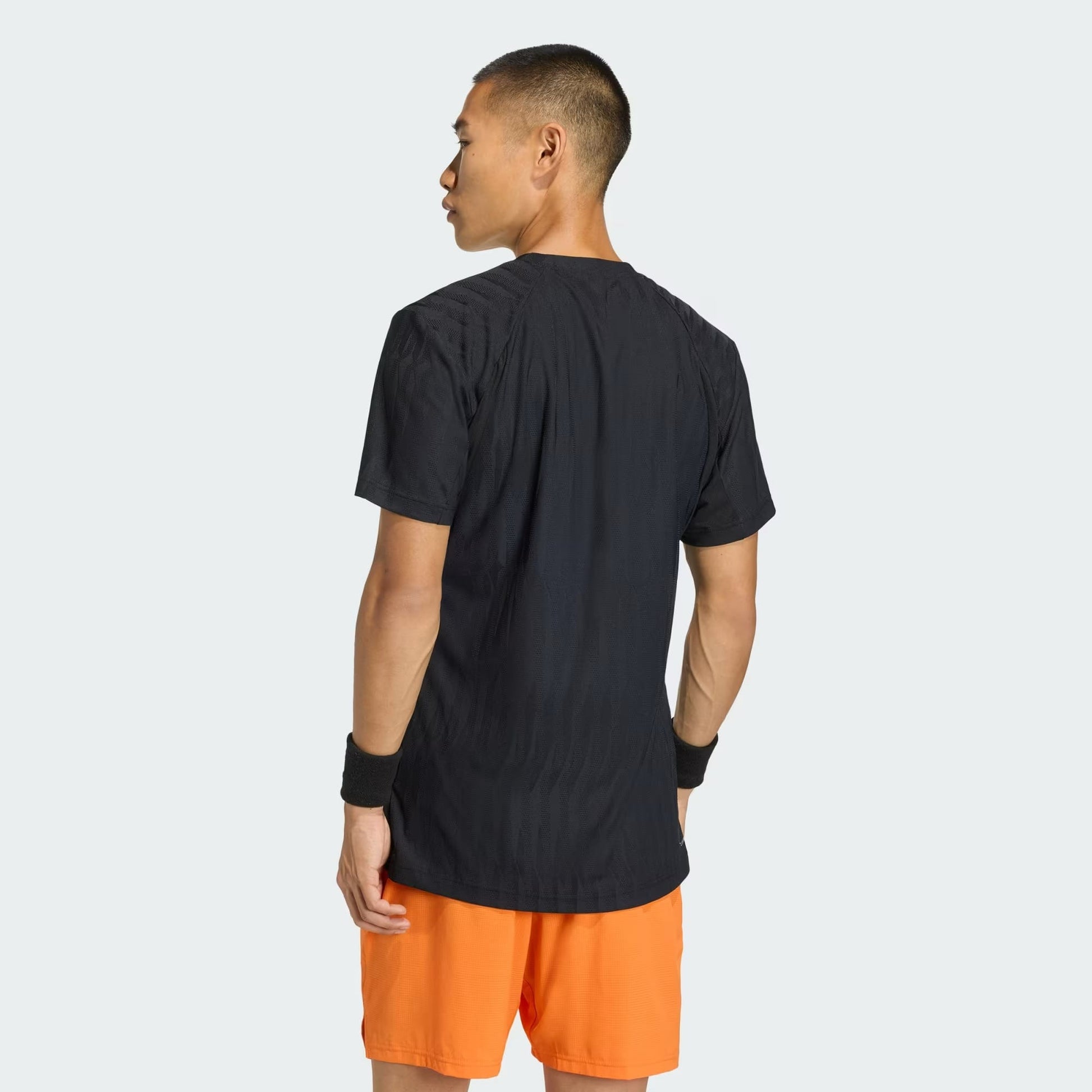 A man stands with his back to the camera, wearing the adidas Climacool+ Airchill Freelift Men's Tennis T-Shirt in black, paired with bright orange shorts and black wristbands, against a plain light background.