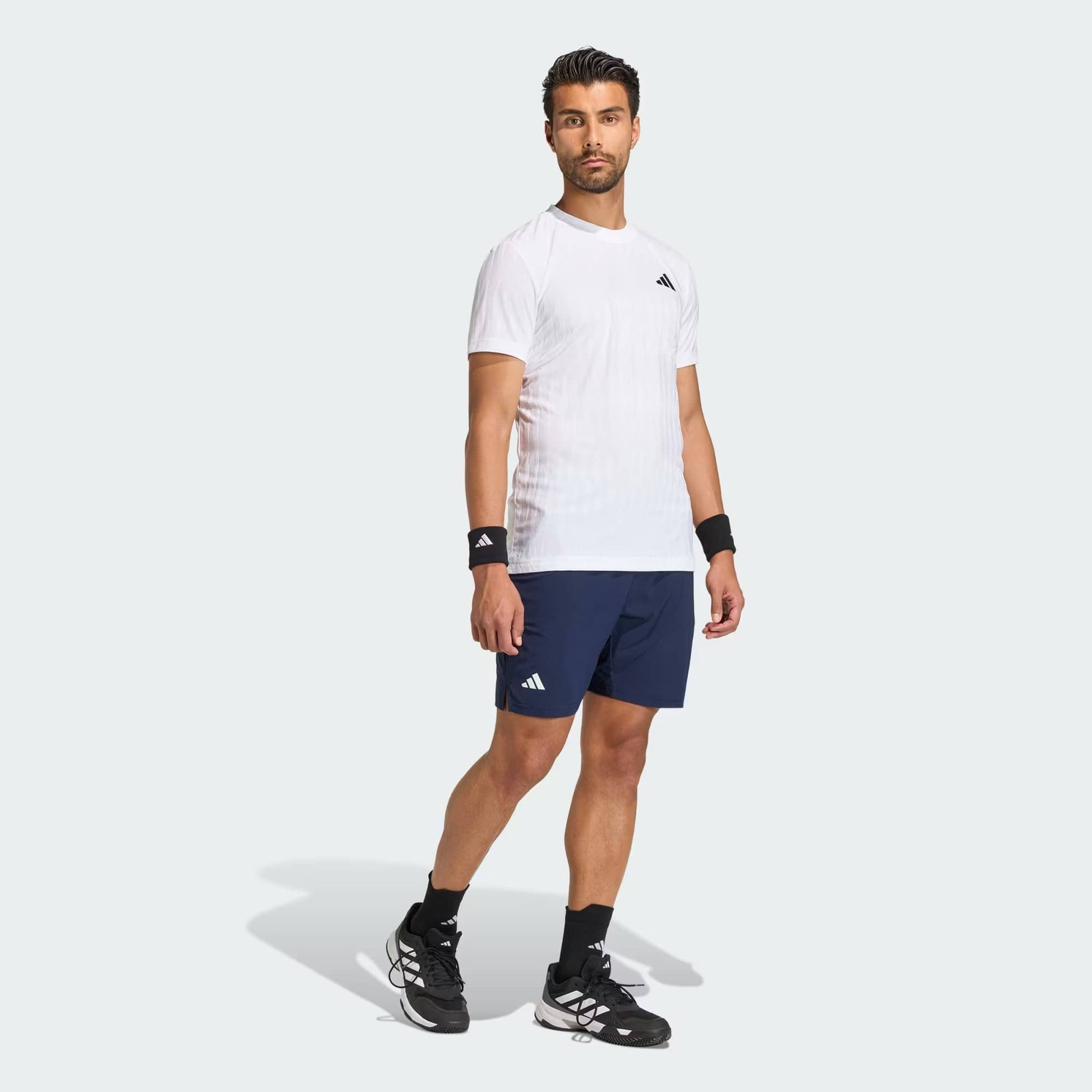 A man stands confidently in an adidas Climacool+ Airchill Freelift Men's Tennis T-Shirt in white, paired with navy adidas shorts, black adidas wristbands, and black athletic shoes against a light gray background.