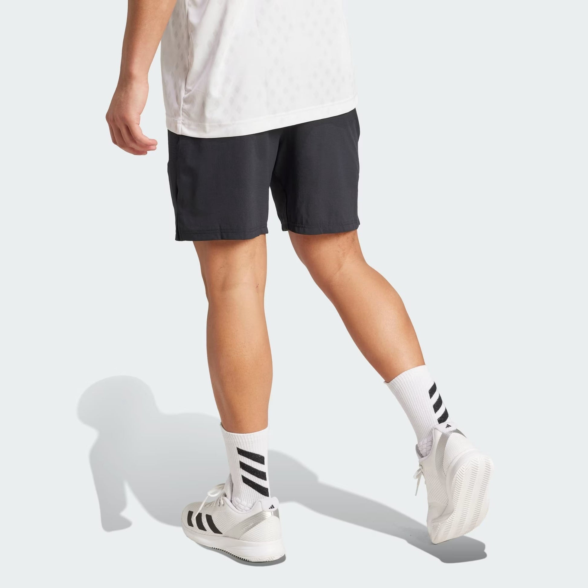 A person is walking away, their lower body visible against a plain light gray background, wearing a white shirt, adidas Climacool Ergo Mens Tennis Shorts in black, white adidas socks with black stripes, and white adidas sneakers.
