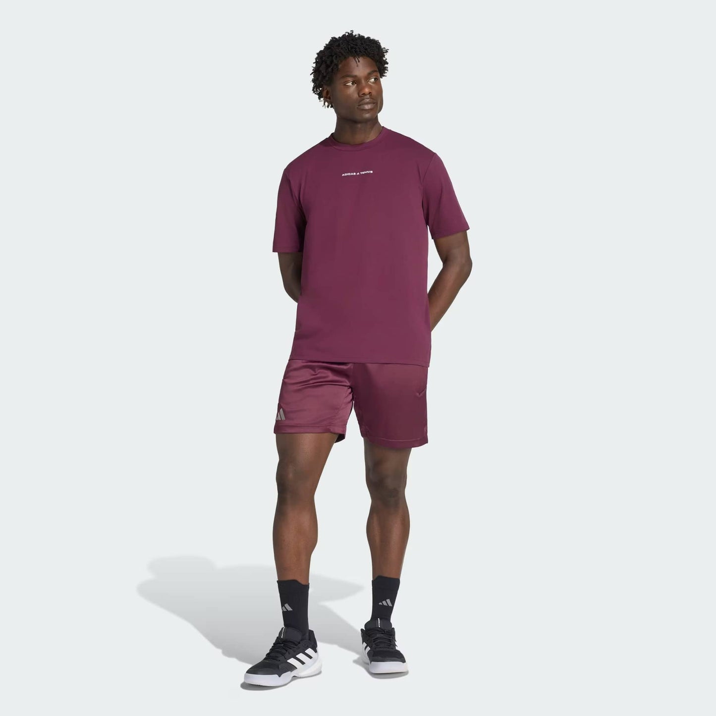 A man stands slightly turned to the side, dressed in a maroon t-shirt and adidas Climacool Ergo Pro Men’s Tennis Shorts in maroon by adidas, paired with black socks and black sneakers, set against a plain light background.