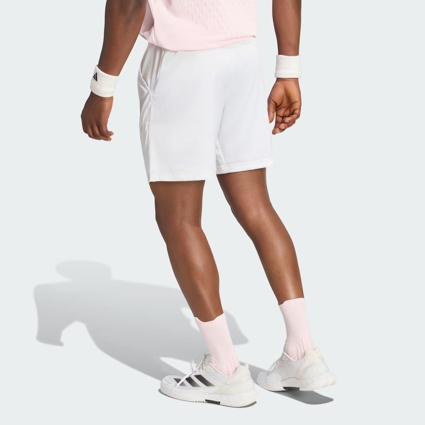 A person wearing a light pink shirt, adidas Climacool Ergo Pro Men’s Tennis Shorts in white, white wristbands, long pink socks, and white adidas sneakers stands on a light gray background, facing away from the camera.