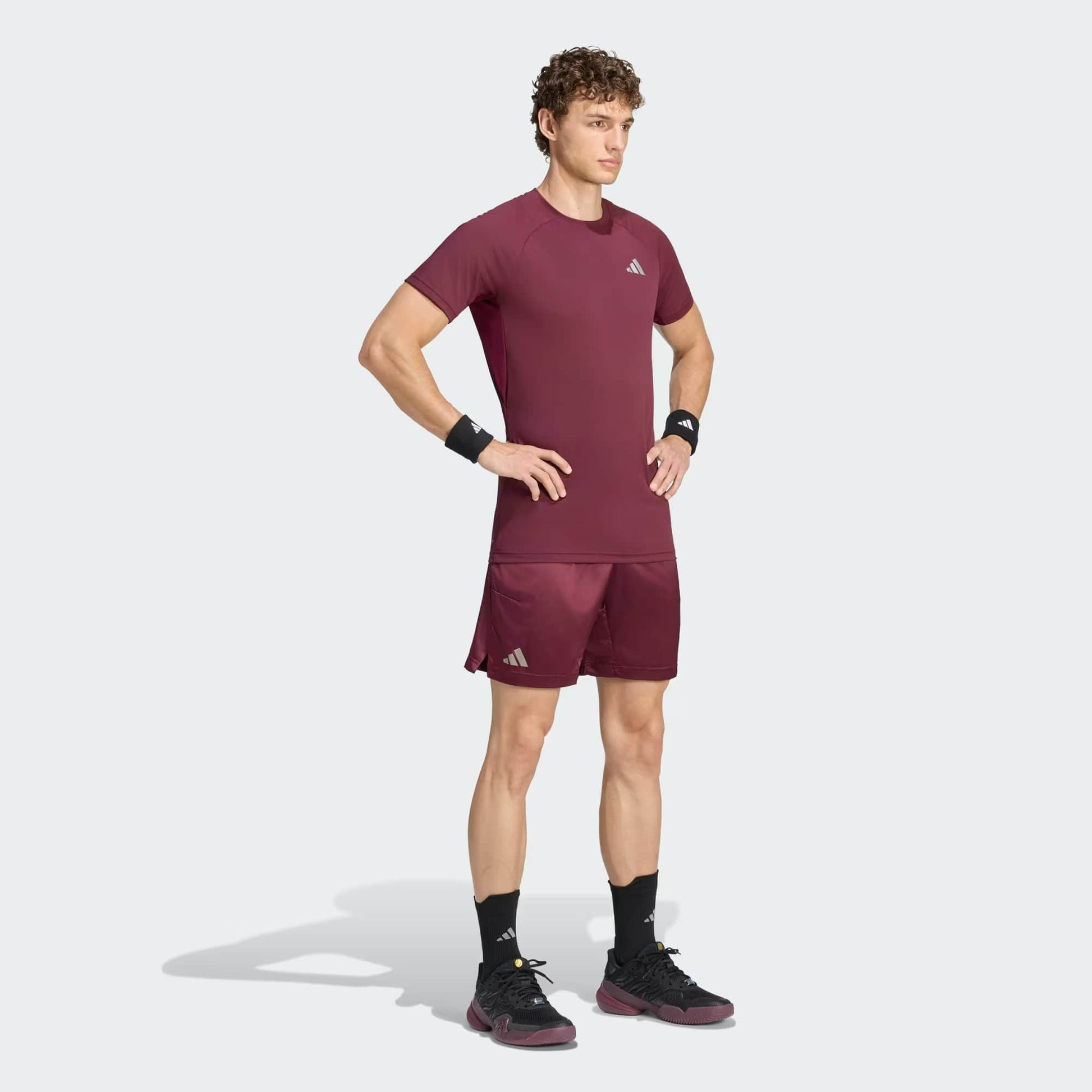 A man stands with his hands on his hips, wearing the adidas Climacool+ Freelift Pro Men’s Tennis T-Shirt in maroon along with matching shorts, black wristbands, socks, and sneakers—all featuring adidas logos—against a plain light background.