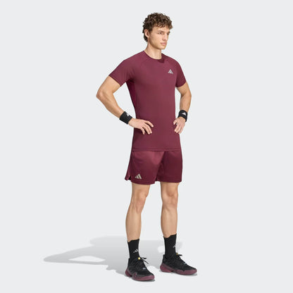 A man stands with his hands on his hips, wearing the adidas Climacool+ Freelift Pro Men’s Tennis T-Shirt in maroon along with matching shorts, black wristbands, socks, and sneakers—all featuring adidas logos—against a plain light background.