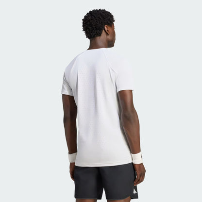 A man stands facing away, wearing the adidas Climacool+ Freelift Pro Men’s Tennis T-Shirt in white, paired with black shorts and white wristbands. The t-shirt features small ventilation holes on the back for breathability. The background is plain and light-colored.