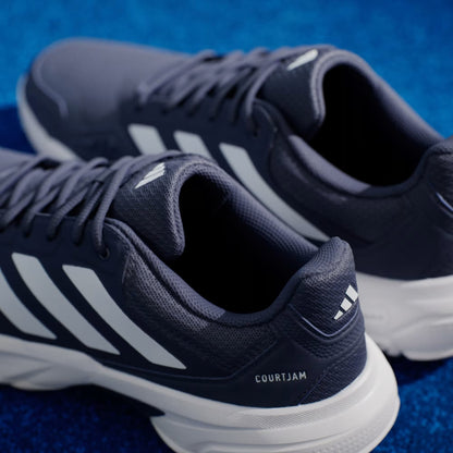 Close-up of the adidas CourtJam Control 3 Men’s Tennis Shoes in Dark Blue and Cloud White, featuring signature white stripes and soles, displayed on a vibrant blue textured surface. Engineered by adidas for optimal court stability, these sneakers offer a perfect blend of style and performance.