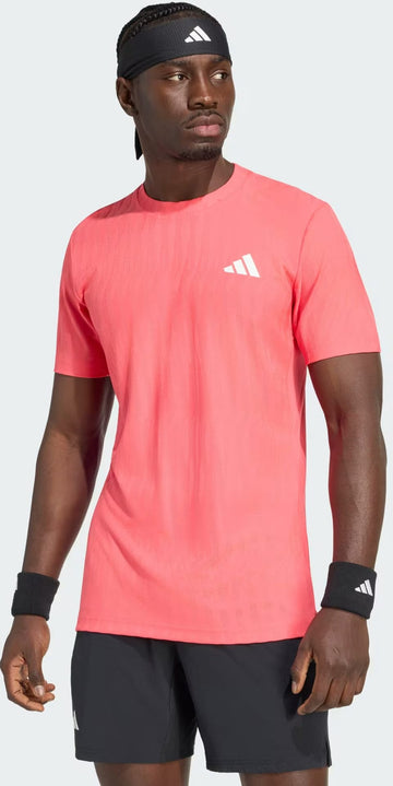 A man wearing an adidas Climacool+ Airchill Freelift Men’s Tennis T-Shirt in Pure Turbo, along with black adidas shorts, a black headband, and black wristbands stands looking to the side against a plain background.