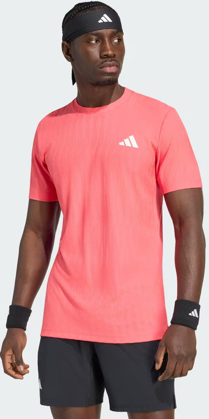A man wearing an adidas Climacool+ Airchill Freelift Men’s Tennis T-Shirt in Pure Turbo, along with black adidas shorts, a black headband, and black wristbands stands looking to the side against a plain background.