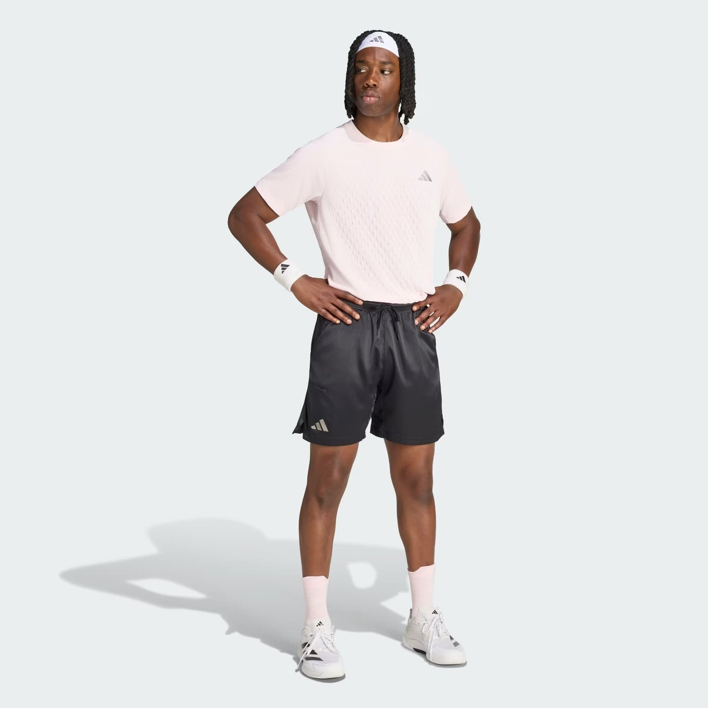 A person stands with hands on hips, wearing a light pink T-shirt, adidas Climacool Ergo Pro Men’s Tennis Shorts in black—designed for quick drying and comfort—white sneakers, pink socks, wristbands, and a white headband. All apparel features the adidas logo and is set against a light gray background.
