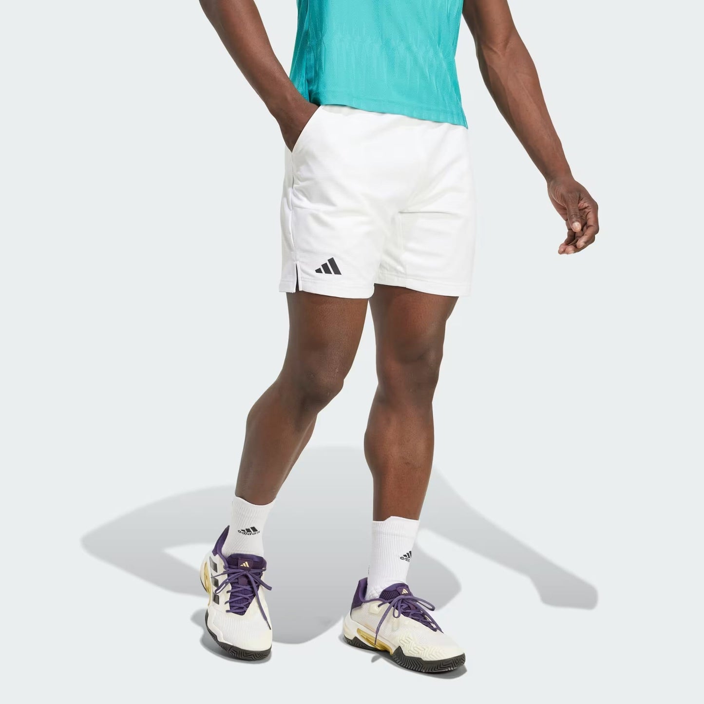 A person in a teal shirt, adidas Climacool Ergo Mens Tennis Shorts (white), white adidas socks, and white sneakers with purple and yellow accents stands against a plain light background.