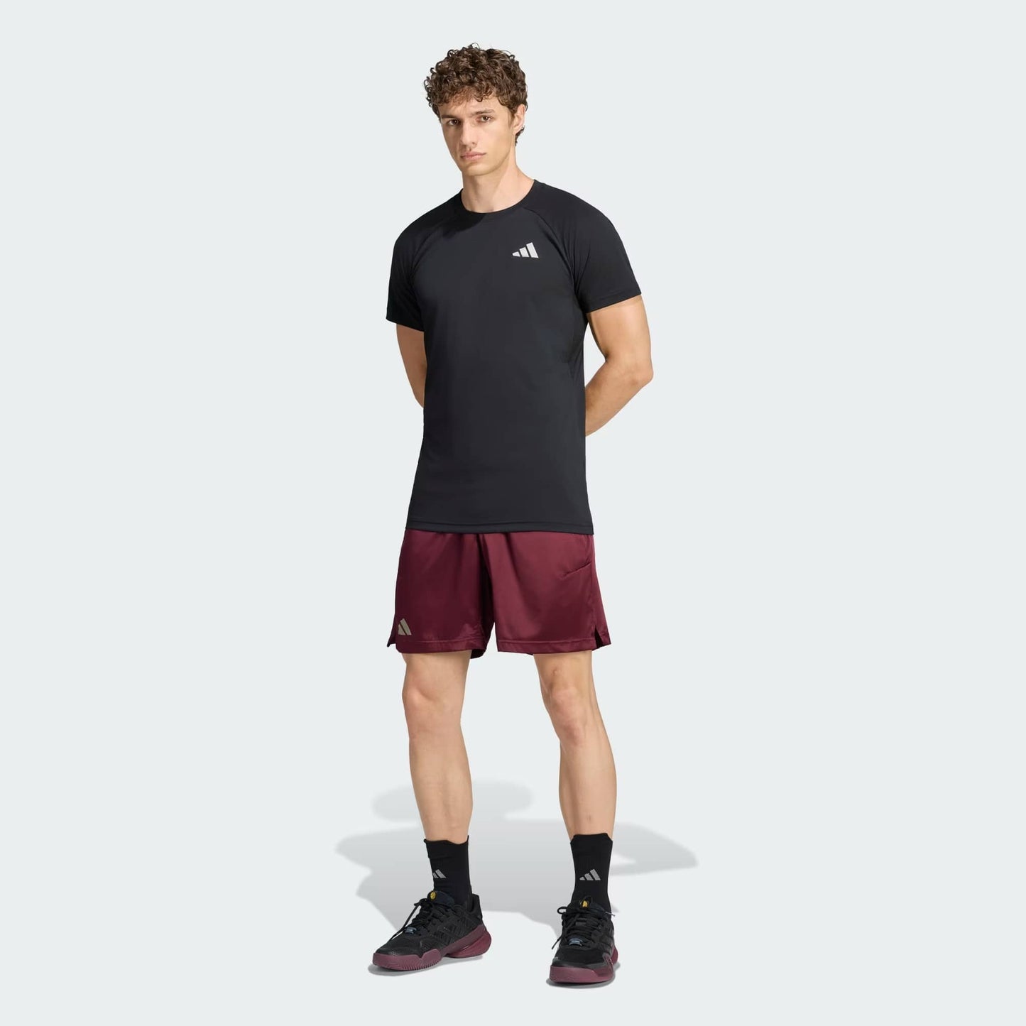 A man stands facing forward wearing the adidas Climacool+ Freelift Pro Men’s Tennis T-Shirt in black, paired with maroon athletic shorts, black socks, and black athletic shoes. The outfit showcases Adidas logos, and the background is plain and light-colored.
