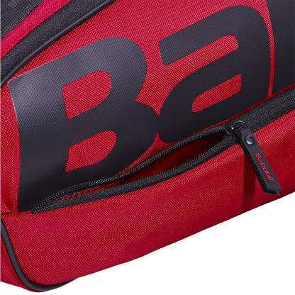 A close-up of the Babolat Court XS 3 Racket Tennis Bag in Burgundy/Black shows a partially unzipped pocket with a zipper pull featuring the "Babolat" brand. Large, bold black "Ba" lettering stands out on the surface of this compact tennis bag.