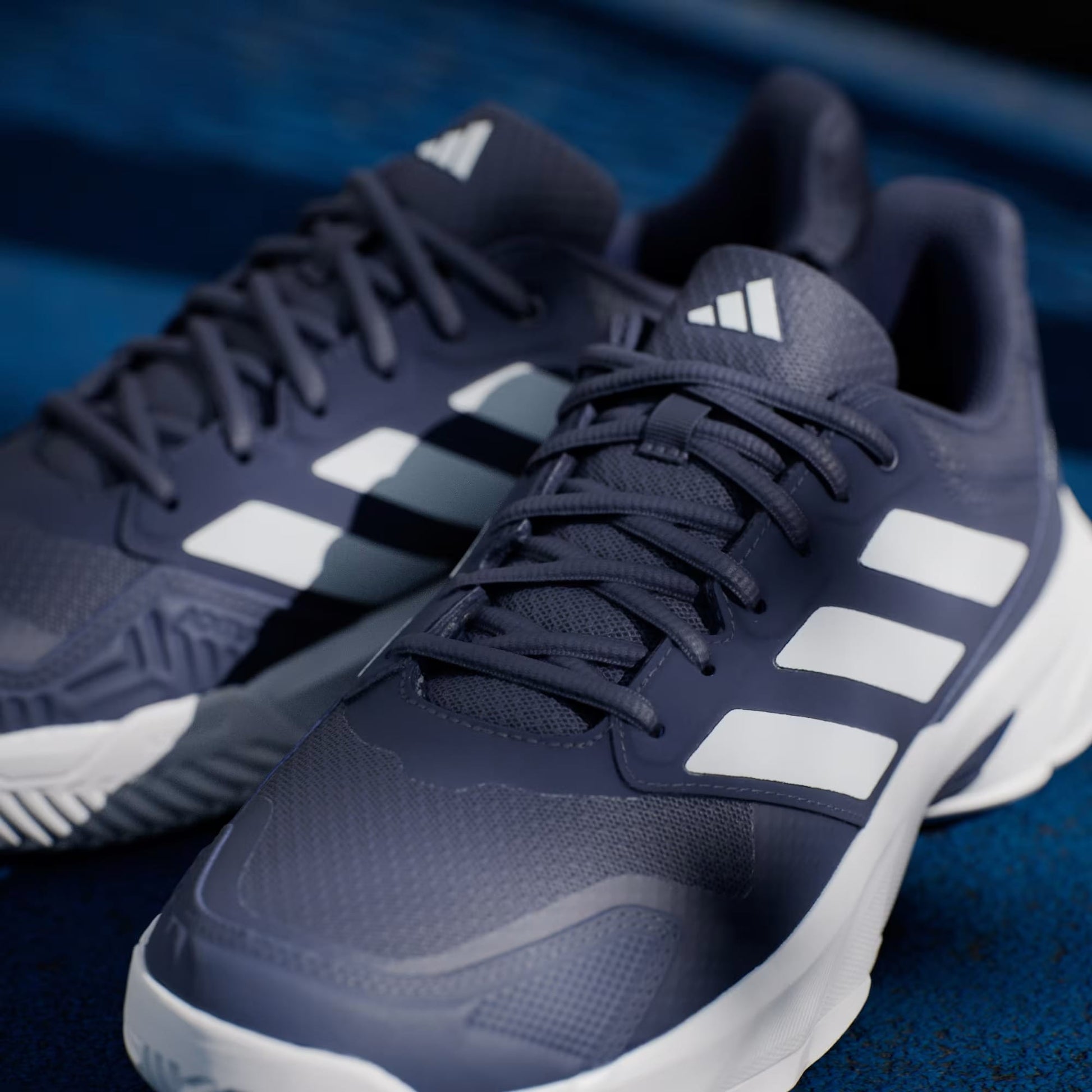 Close-up of adidas CourtJam Control 3 Men’s Tennis Shoes in Dark Blue and Cloud White, featuring signature white stripes and white soles, displayed on a blue textured surface for enhanced stability.