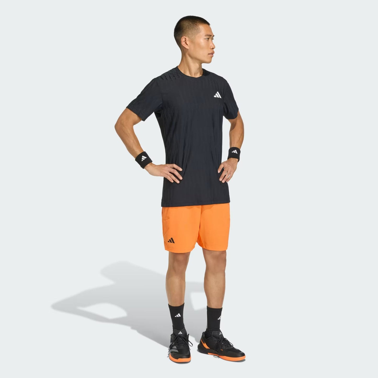 A man stands with hands on hips, looking to the side, wearing a black adidas Climacool+ Airchill Freelift Men's Tennis T-Shirt against a plain light background.