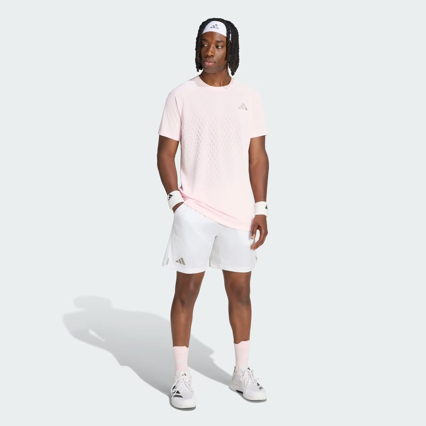 A man wearing a light pink t-shirt, white adidas Climacool Ergo Pro Men’s Tennis Shorts, white sneakers, and a white headband stands facing slightly right against a plain background. He has wristbands on both arms and his hands at his sides.