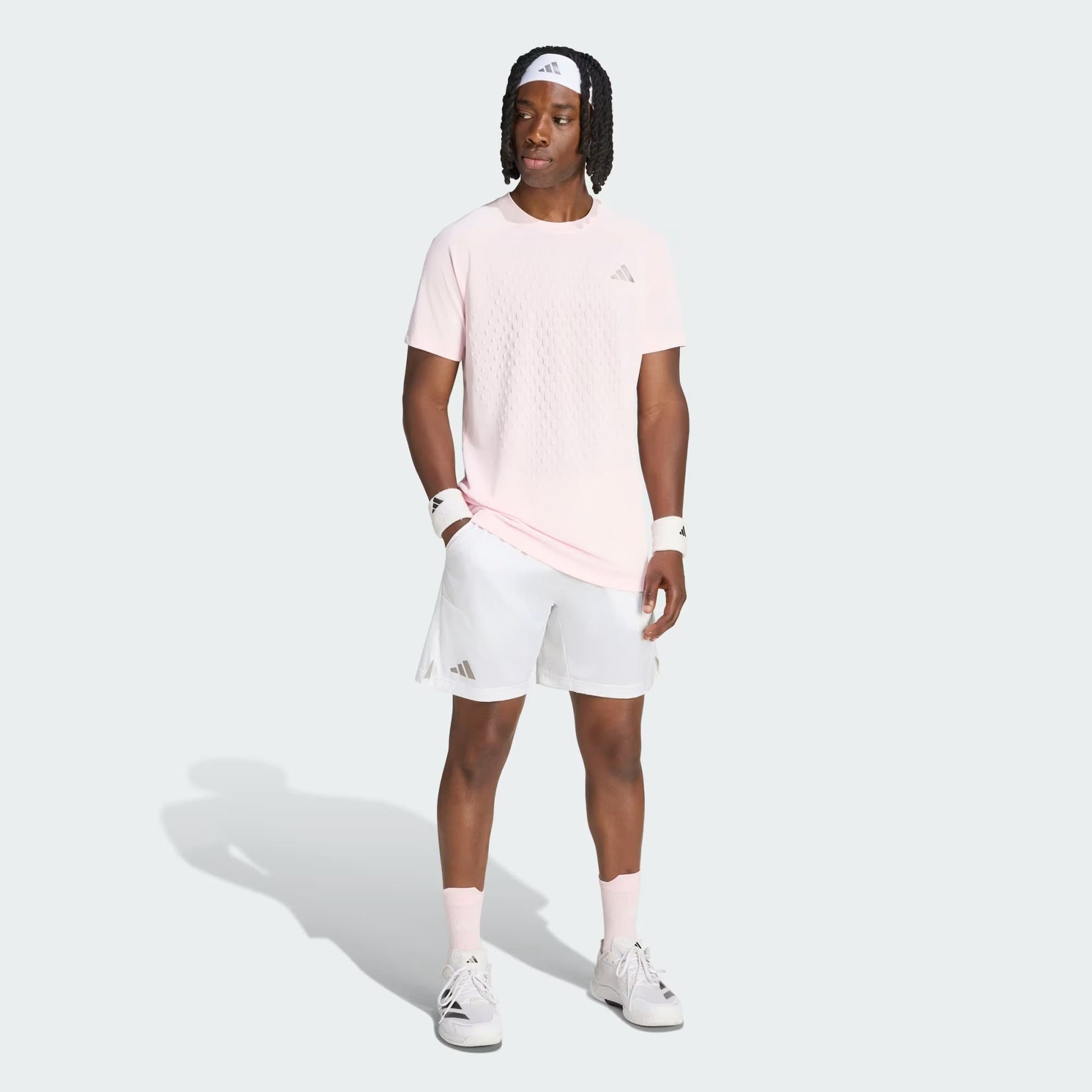 A man wearing a light pink t-shirt, white adidas Climacool Ergo Pro Men’s Tennis Shorts, white sneakers, and a white headband stands facing slightly right against a plain background. He has wristbands on both arms and his hands at his sides.