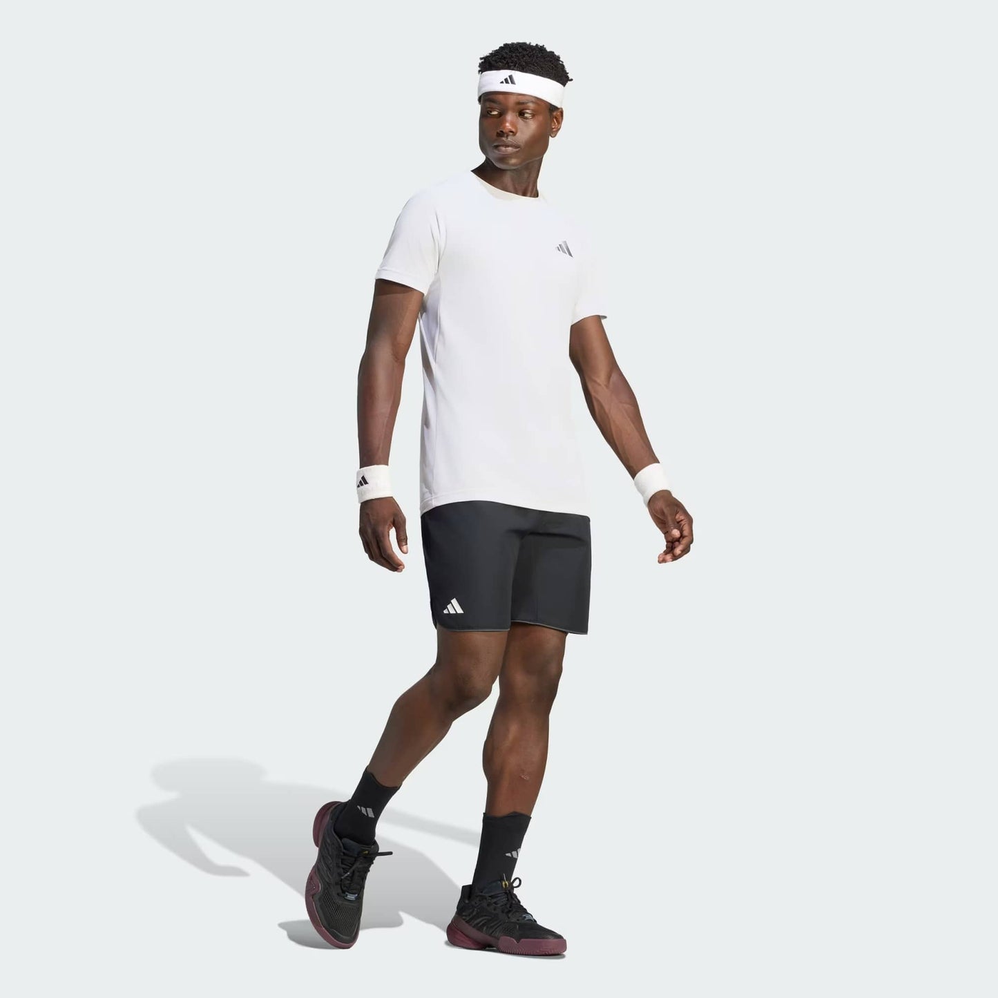 A man wearing an adidas Climacool+ Freelift Pro Men’s Tennis T-Shirt in white, black shorts, black sneakers, a white headband, and white wristbands stands and looks to the side against a light background.