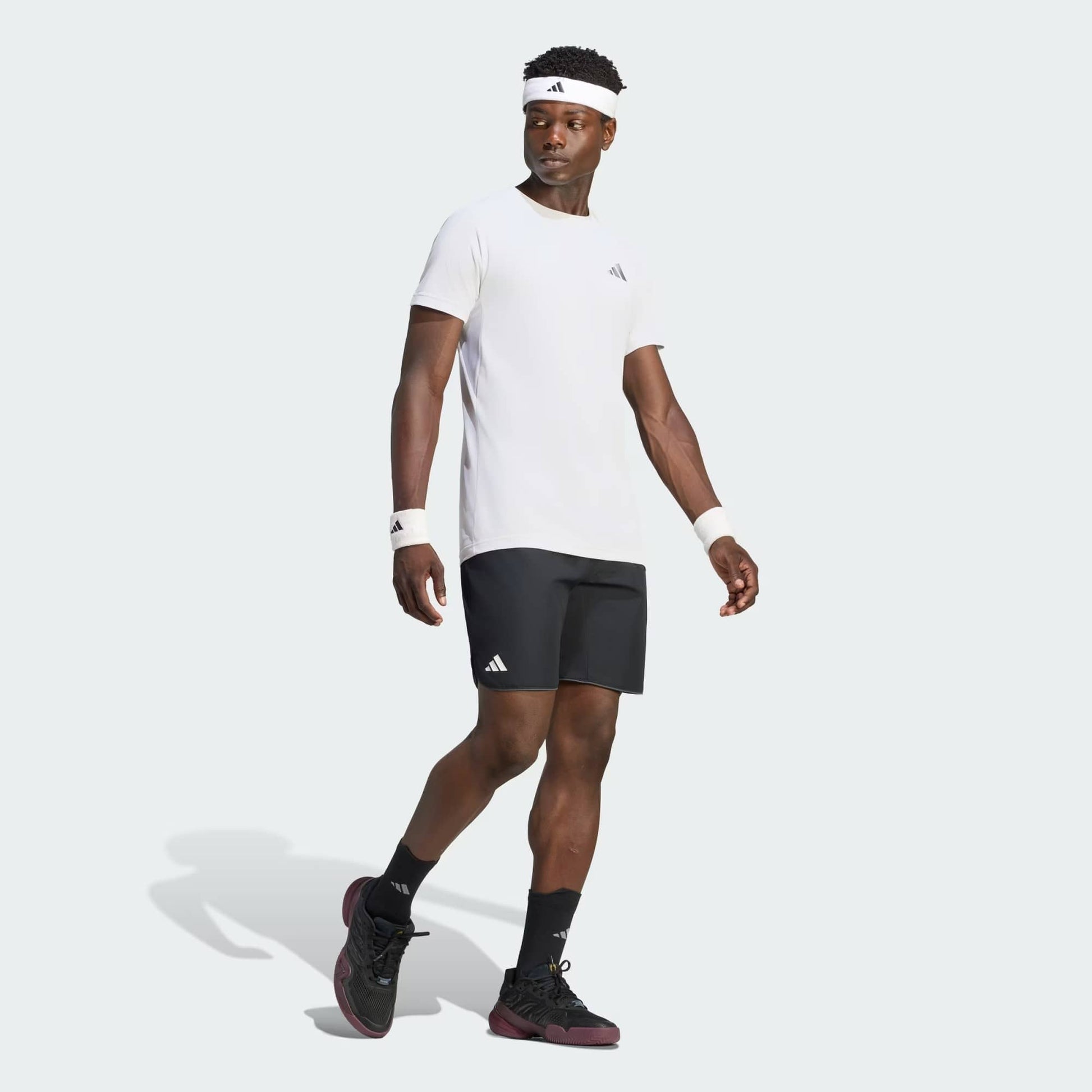 A man wearing an adidas Climacool+ Freelift Pro Men’s Tennis T-Shirt in white, black shorts, black sneakers, a white headband, and white wristbands stands and looks to the side against a light background.