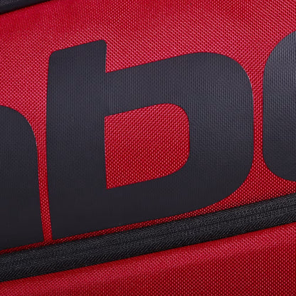 Close-up of the Babolat Court XS 3 Racket Tennis Bag in Burgundy and Black, showcasing its red textured fabric and large black brand lettering. A black zipper runs diagonally across the lower part of the image, highlighting this compact Babolat tennis bag.