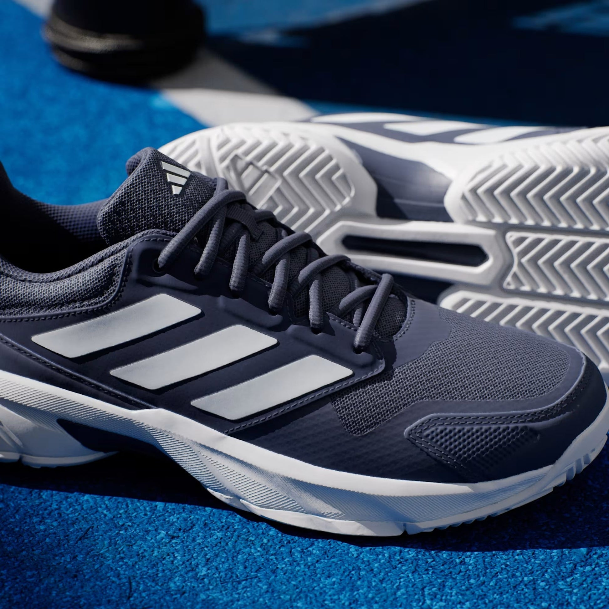 A close-up of the adidas CourtJam Control 3 Men’s Tennis Shoes in Dark Blue and Cloud White, featuring signature white adidas stripes, is displayed on a blue track. One shoe stands upright while the other lies on its side to showcase the patterned tread designed for enhanced stability.
