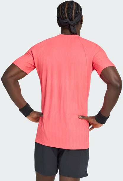 A man with braided hair and a headband faces away, wearing a bright pink adidas Climacool+ Airchill Freelift Men’s Tennis T-Shirt in Pure Turbo, paired with black wristbands and black shorts against a plain light background.