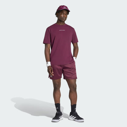 A man wearing matching maroon adidas Climacool Ergo Pro Men’s Tennis Shorts, a burgundy T-shirt, and a cap, paired with black and white adidas sneakers, black socks, and white wristbands, stands against a light background.