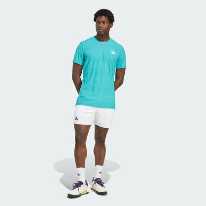 A man stands against a light background wearing a turquoise adidas t-shirt, adidas Climacool Ergo Men's Tennis Shorts in white, white socks, and white athletic shoes with black accents. His hands are behind his back as he looks slightly to the side.