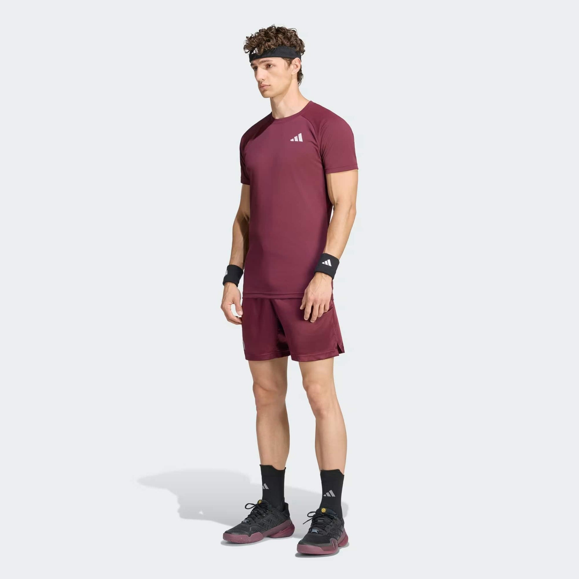 A man stands facing forward against a plain light background, dressed in a coordinated maroon athletic ensemble. He wears an adidas Climacool+ Freelift Pro Men’s Tennis T-Shirt in maroon, paired with matching shorts, black socks, sneakers, wristbands, and a headband.