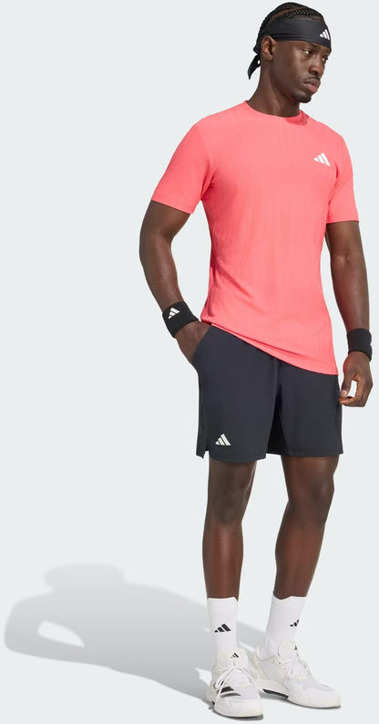A man wearing an adidas Climacool+ Airchill Freelift Men’s Tennis T-Shirt in Pure Turbo, paired with black shorts, white sneakers, white socks, and black wristbands stands against a plain background.