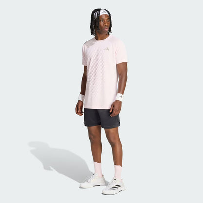 A man stands against a plain light background, wearing a light pink adidas shirt, adidas Climacool Ergo Pro Men’s Tennis Shorts in black, white wristbands, pink socks, and white sneakers. He looks to the side with his arms relaxed by his sides.