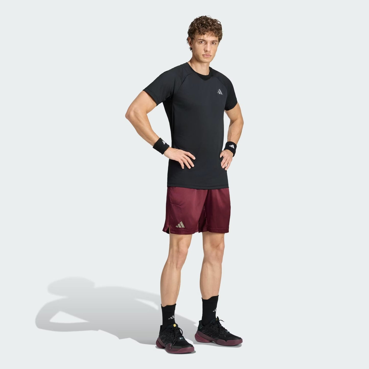 A man stands with hands on hips, wearing the adidas Climacool+ Freelift Pro Men’s Tennis T-Shirt in black, paired with maroon athletic shorts, black wristbands, black socks, and black sneakers against a light gray background.