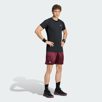 A man stands with hands on hips, wearing the adidas Climacool+ Freelift Pro Men’s Tennis T-Shirt in black, paired with maroon athletic shorts, black wristbands, black socks, and black sneakers against a light gray background.