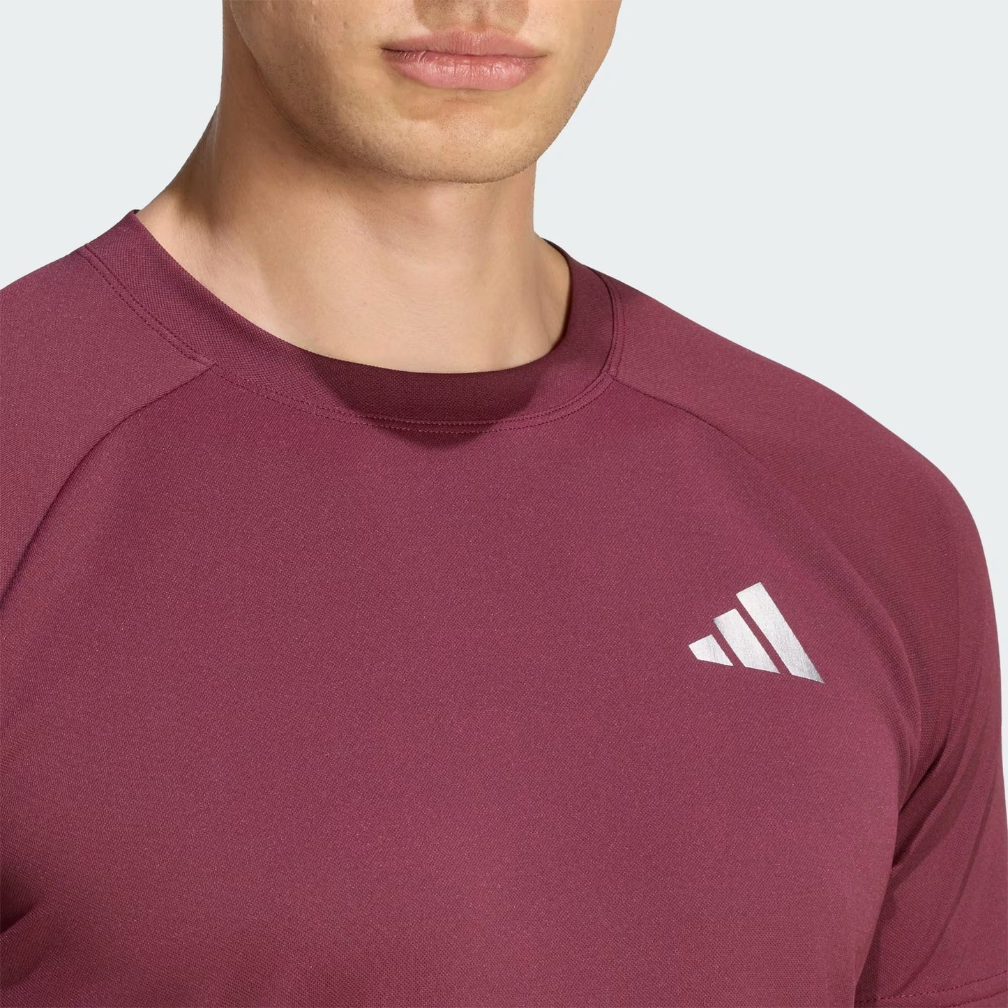 A close-up of a person wearing the adidas Climacool+ Freelift Pro Men’s Tennis T-Shirt in maroon, featuring a white adidas logo on the chest. Only the lower half of the face and upper torso are visible.