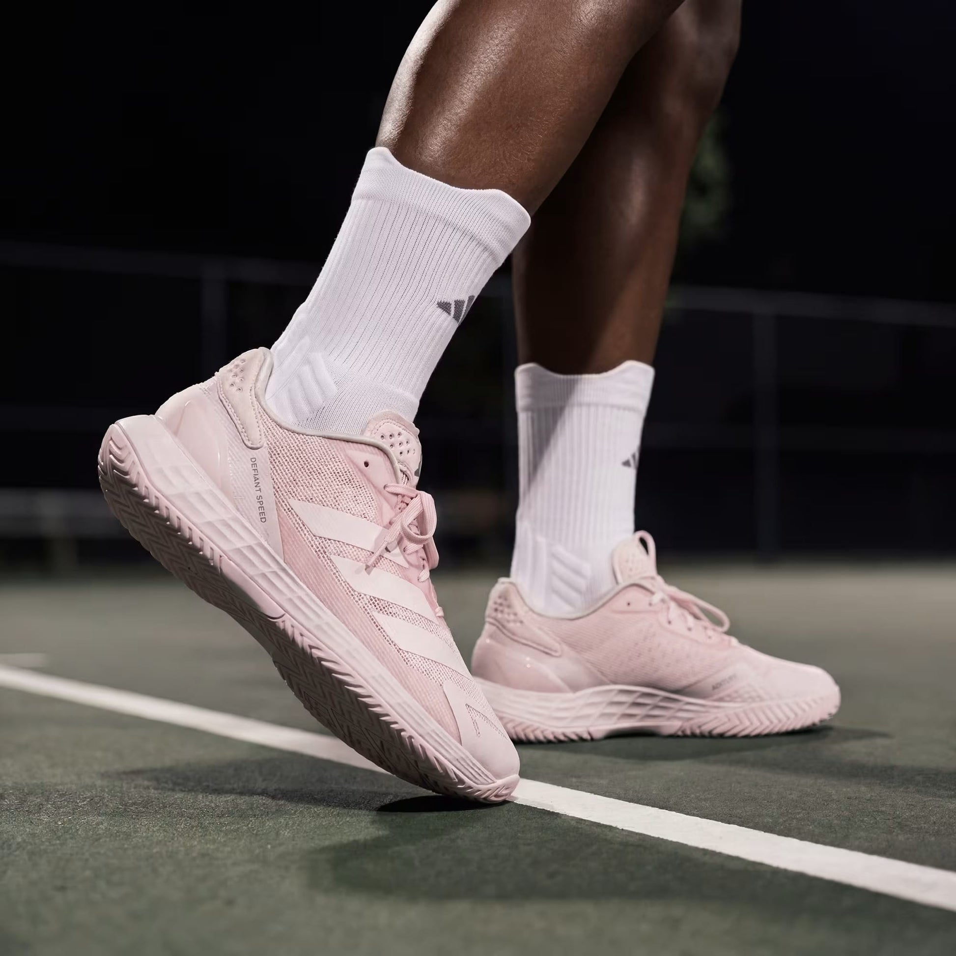 A person wearing adidas Defiant Speed 2 Womens Tennis Shoes in Distilled Pink and white adidas socks stands on a tennis court at night, poised to move. The lighting emphasizes the lightweight shoes and lower legs, while the background remains blurred.