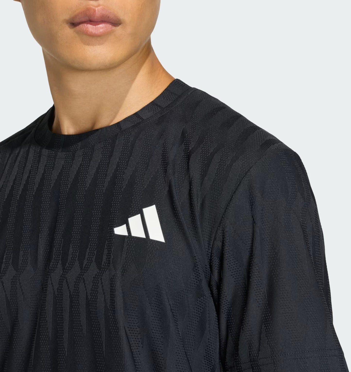 A man wears the adidas Climacool+ Airchill Freelift Mens Tennis T-Shirt in black, featuring a subtle pattern and white adidas logo on the chest, shown from shoulders up against a plain light background. Moisture-wicking fabric keeps him cool and comfortable.
