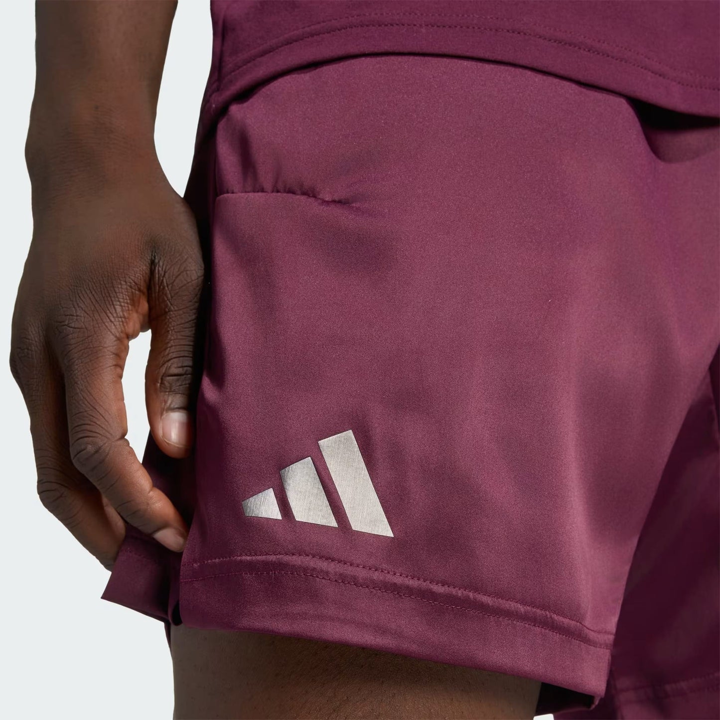 A close-up of a person wearing adidas Climacool Ergo Pro Mens Tennis Shorts in maroon, featuring a white adidas logo on the left leg. The person’s hand is positioned near the ergonomic side seam of the shorts.