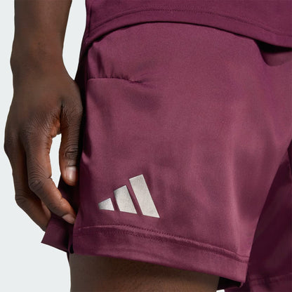 A close-up of a person wearing adidas Climacool Ergo Pro Mens Tennis Shorts in maroon, featuring a white adidas logo on the left leg. The person’s hand is positioned near the ergonomic side seam of the shorts.