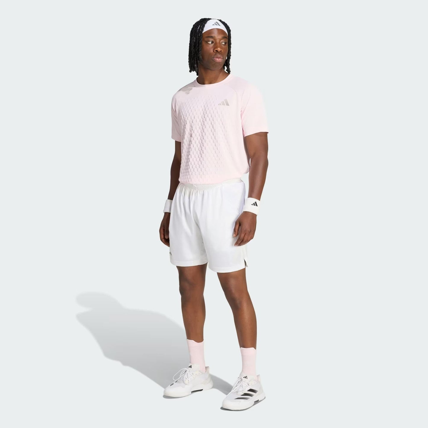 A man wearing a light pink adidas shirt, adidas Climacool Ergo Pro Mens Tennis Shorts in white, white adidas socks and shoes, and a white headband stands on a light gray background.