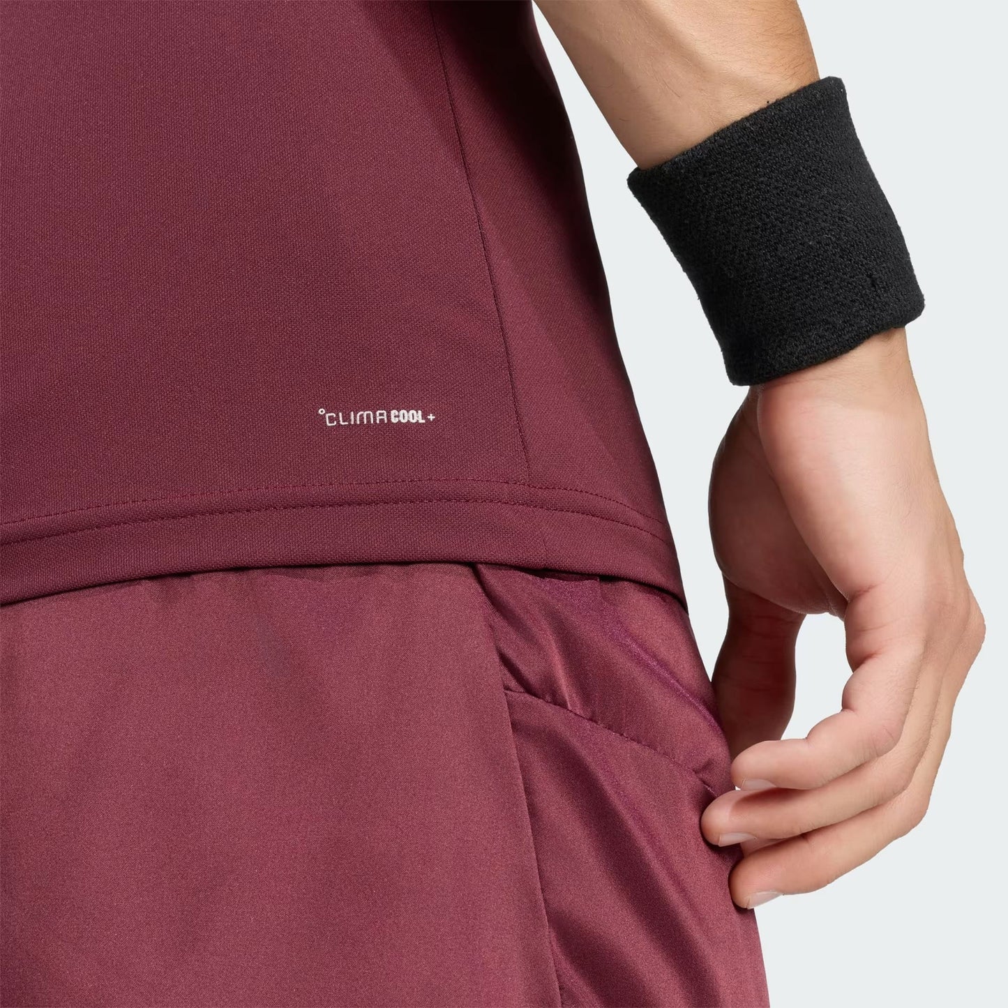 A person wearing the adidas Climacool+ Freelift Pro Men’s Tennis T-Shirt in maroon and matching maroon athletic shorts, resting their hand near the pocket. They complete the effortless adidas Pro tennis style with a black wristband.