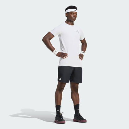 A man wearing an adidas Climacool+ Freelift Pro Men’s Tennis T-Shirt in white, paired with black shorts, wristbands, a headband, socks, and sneakers stands with his hands on his hips against a plain background.