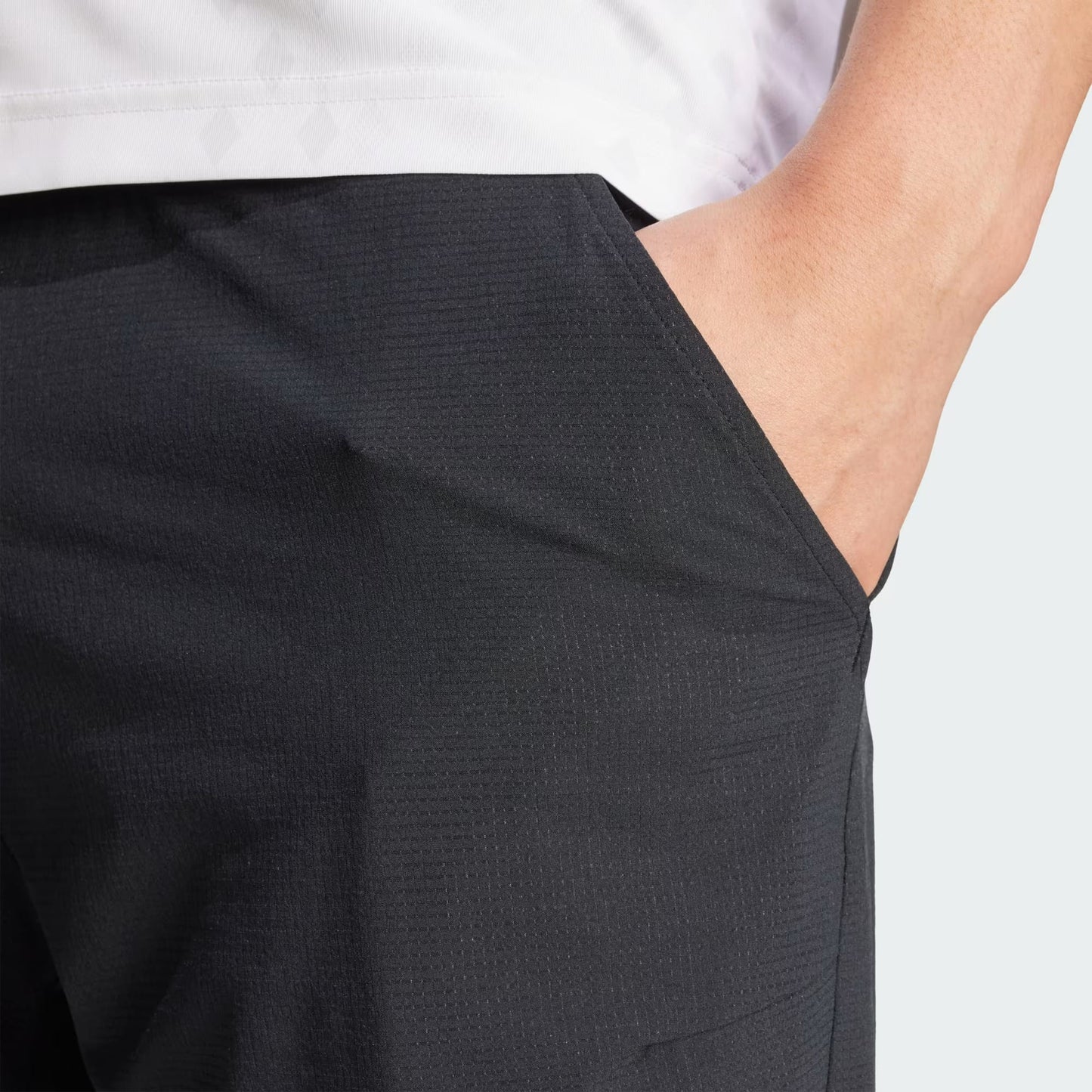 Close-up of a hand in the pocket of adidas Climacool Ergo Mens Tennis Shorts in black. Only the lower torso and upper thigh are visible, with a white shirt and plain light gray background highlighting the sweat-wicking fabric.