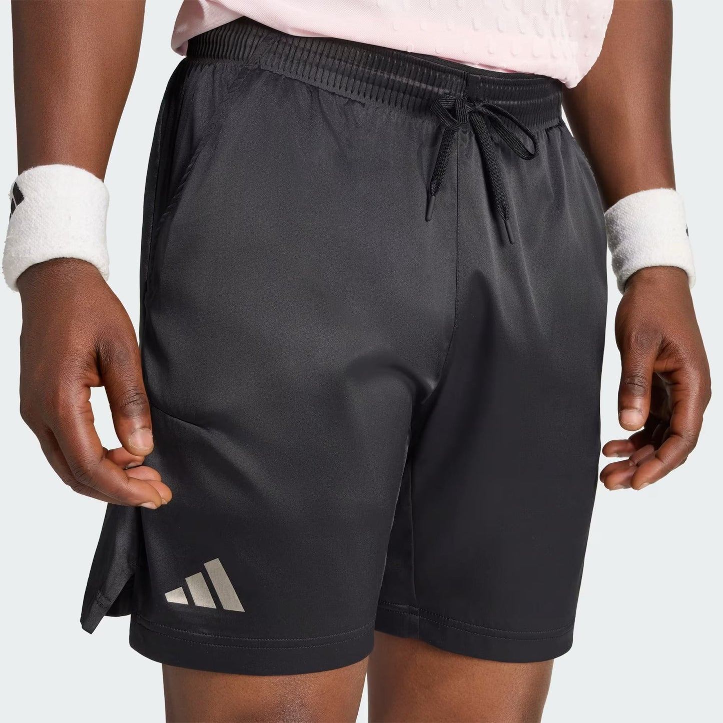A person is shown wearing adidas Climacool Ergo Pro Men’s Tennis Shorts in black, paired with a light pink textured shirt and white wristbands. Only the lower torso, hands, and upper thighs are visible. The adidas logo appears on the left leg of these quick-drying tennis shorts.