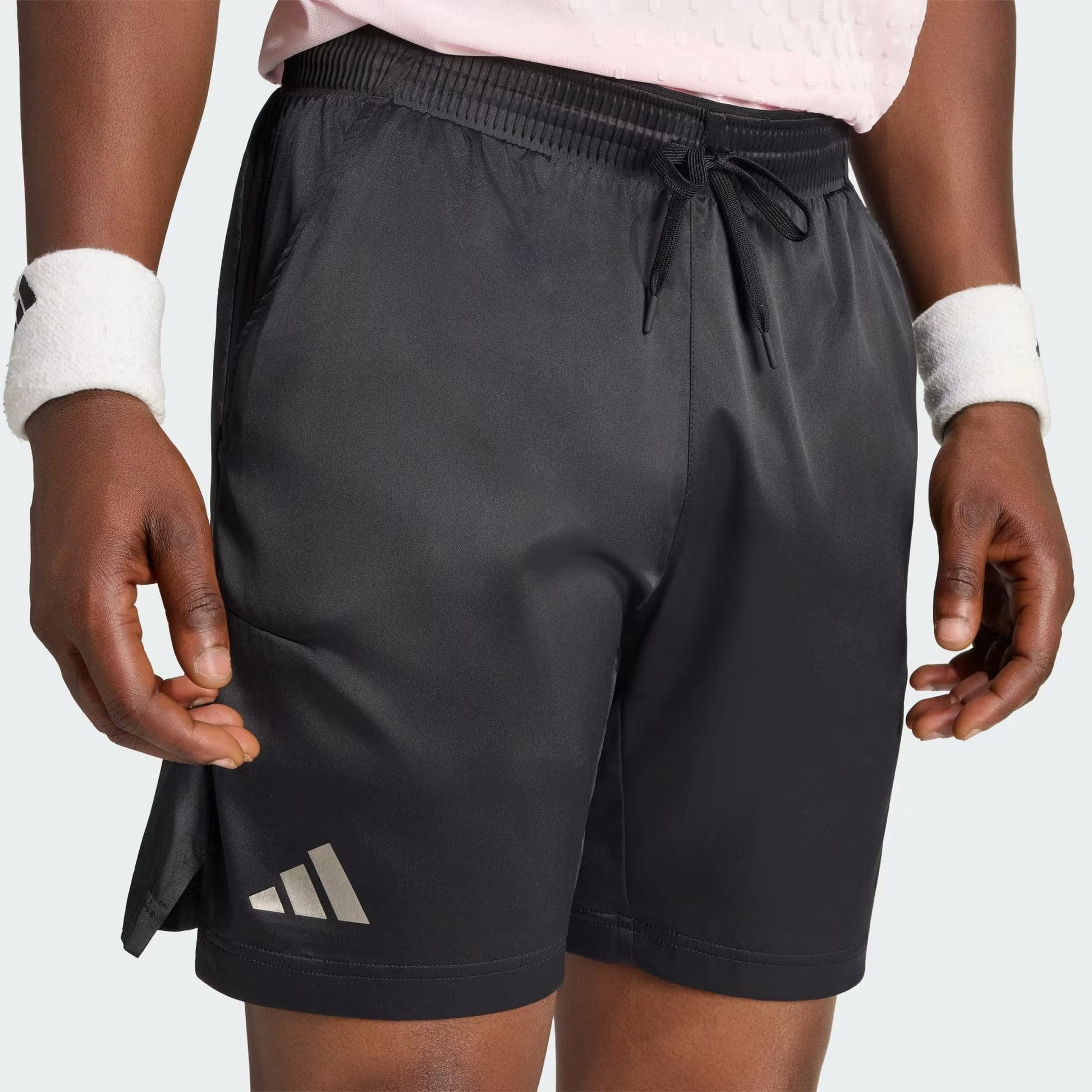 A person is shown wearing adidas Climacool Ergo Pro Men’s Tennis Shorts in black, paired with a light pink textured shirt and white wristbands. Only the lower torso, hands, and upper thighs are visible. The adidas logo appears on the left leg of these quick-drying tennis shorts.