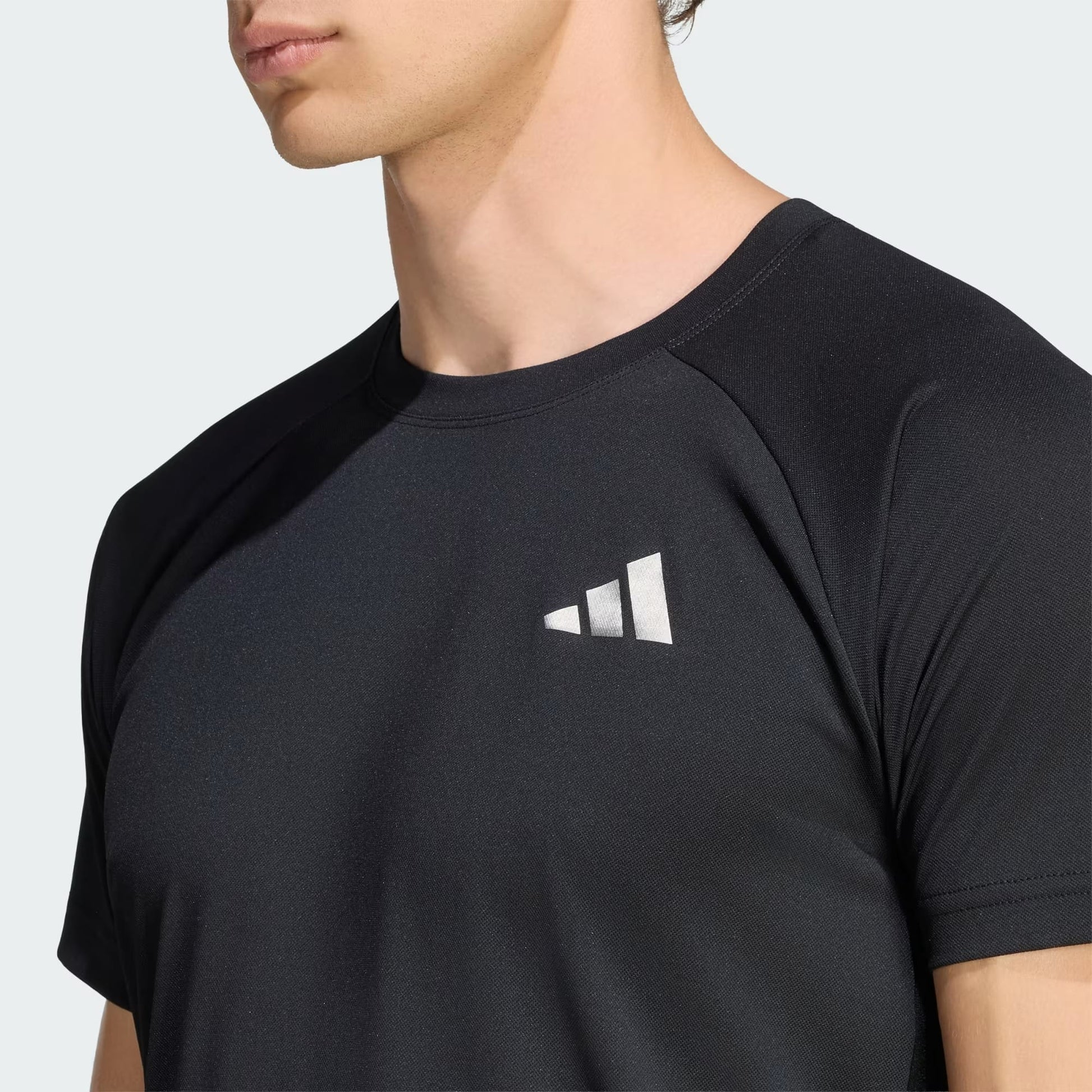 A man is shown from the shoulders up against a plain light background, wearing the adidas Climacool+ Freelift Pro Men’s Tennis T-Shirt in black, which features a white geometric adidas logo on the chest.