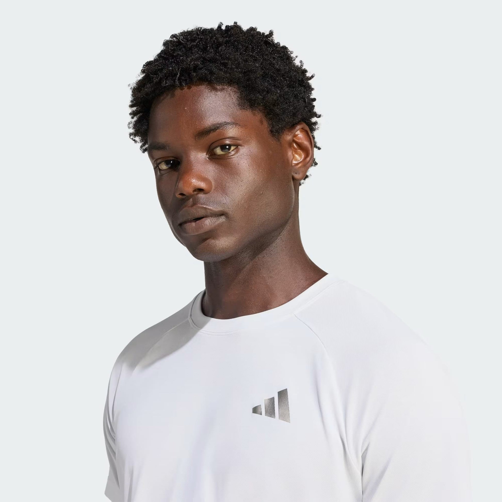 A person with short curly hair, wearing the adidas Climacool+ Freelift Pro Men’s Tennis T-Shirt in white, which features a small geometric logo, stands against a plain light background and looks directly at the camera with a neutral expression.
