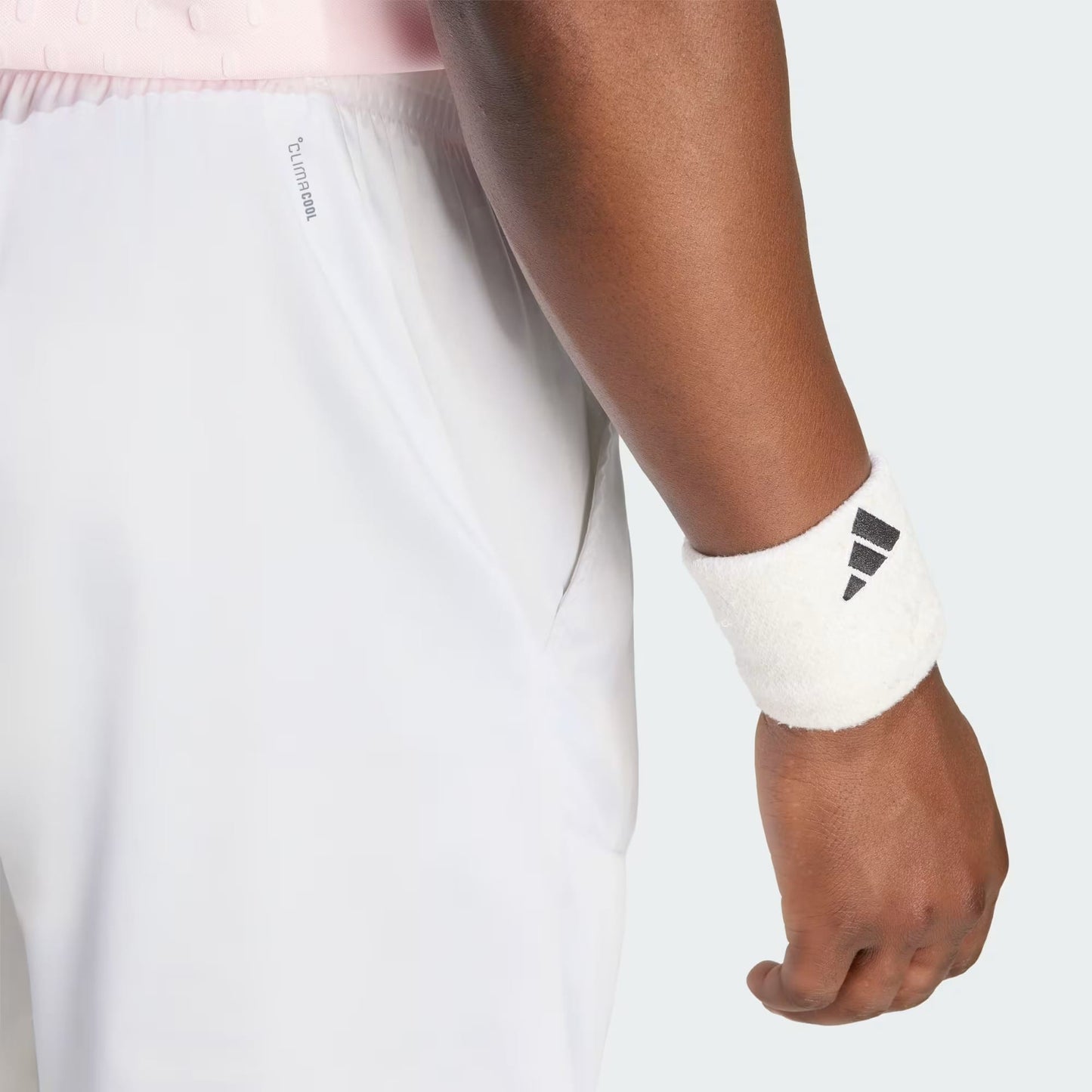 A close-up of a person wearing adidas Climacool Ergo Pro Men’s Tennis Shorts in white and a white adidas wristband with a black logo on their right wrist, standing against a plain background.