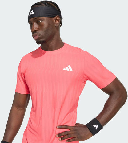 A man wearing an adidas Climacool+ Airchill Freelift Men’s Tennis T-Shirt in Pure Turbo, paired with a black headband and wristbands, stands with one hand on his hip, gazing to the side against a plain background.