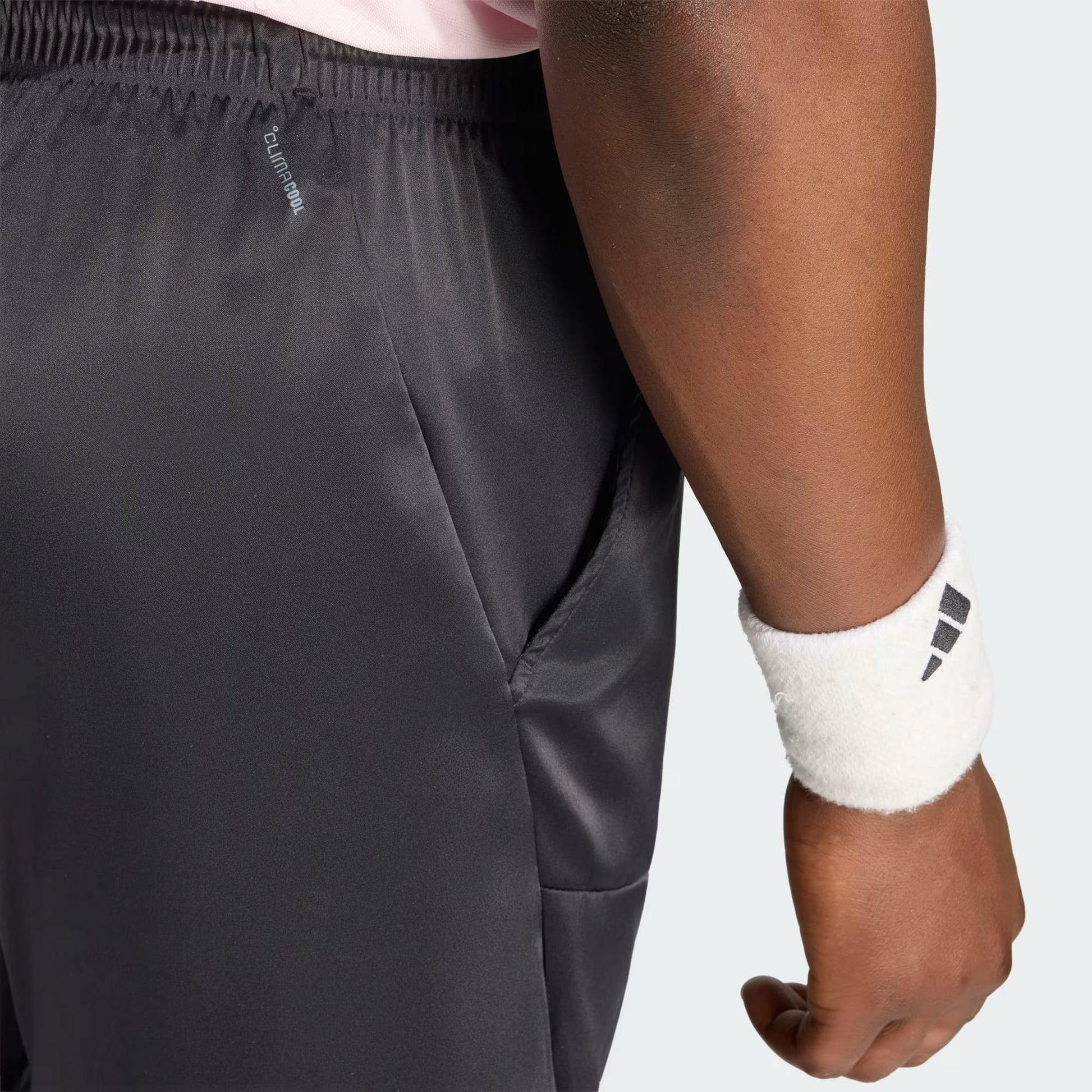 A person shown from the waist down wearing adidas Climacool Ergo Pro Men’s Tennis Shorts in black and a white wristband featuring the black adidas logo, with their hand resting by their side.