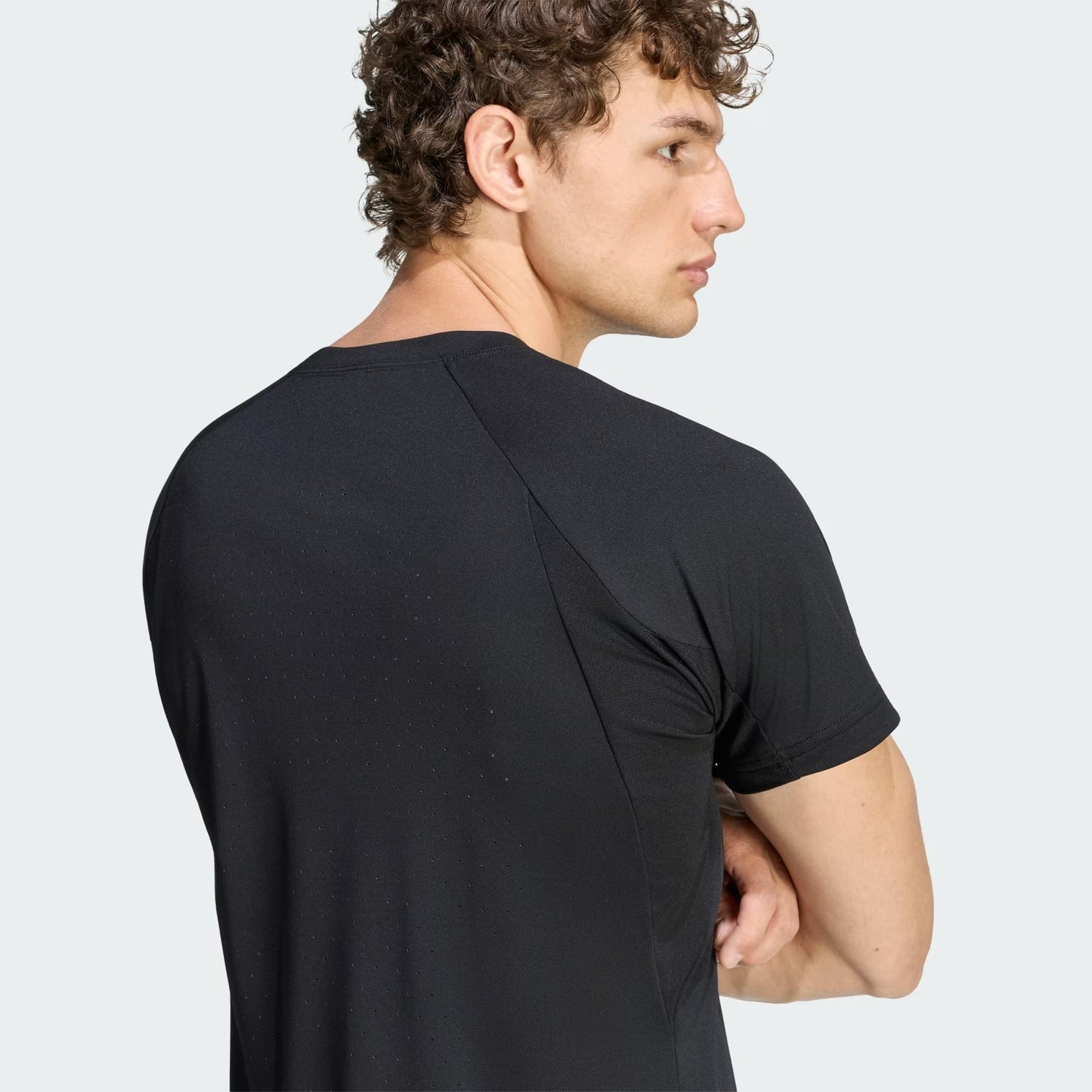 A man with curly hair, wearing the adidas Climacool+ Freelift Pro Men’s Tennis T-Shirt in black, stands with his arms crossed, facing away and looking to the side.