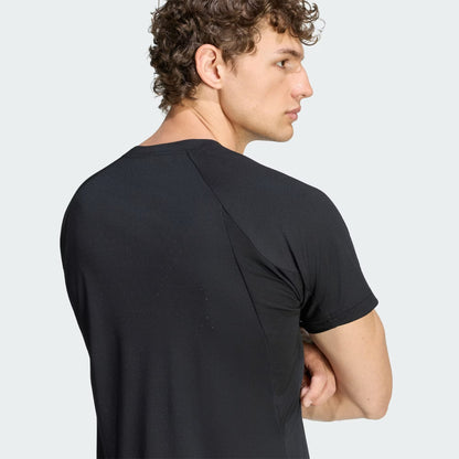 A man with curly hair, wearing the adidas Climacool+ Freelift Pro Men’s Tennis T-Shirt in black, stands with his arms crossed, facing away and looking to the side.